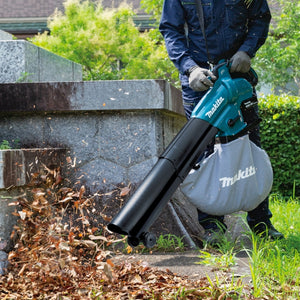 Person using a Makita leaf blower to clear leaves from a garden area.